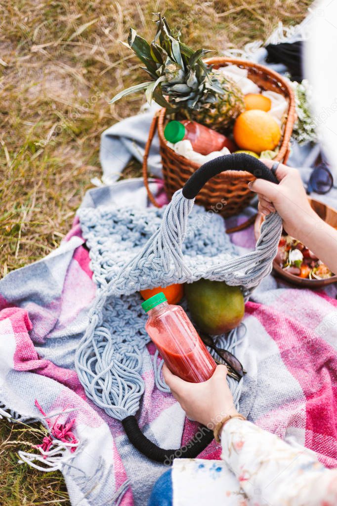 Close up photo of woman hand holding juice bottle. Basket full of fruits on picnic blanket in park