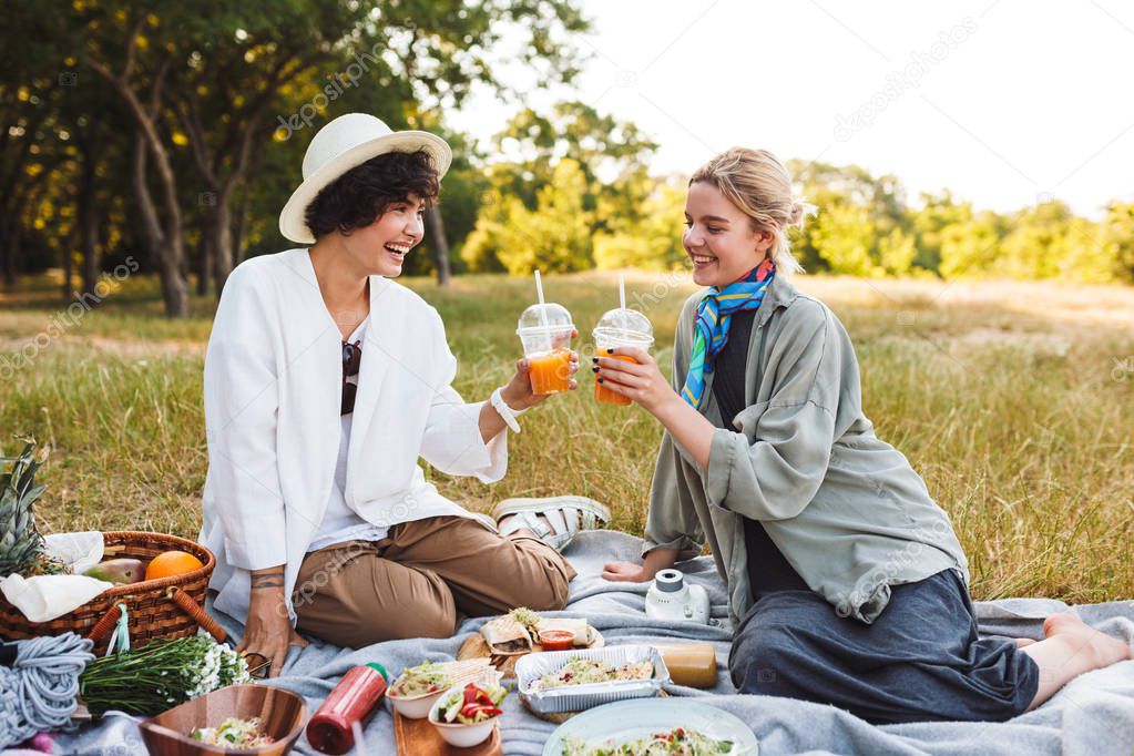 Two pretty smiling girls sitting on picnic blanket holding orange juice in hands happily spending time on picnin in park