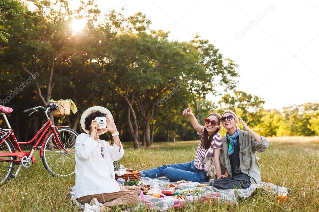 Beautiful girls sitting on picnic blanket with little dog and happily taking photos on white polaroid camera spending time on picnic in park with red bicycle on background