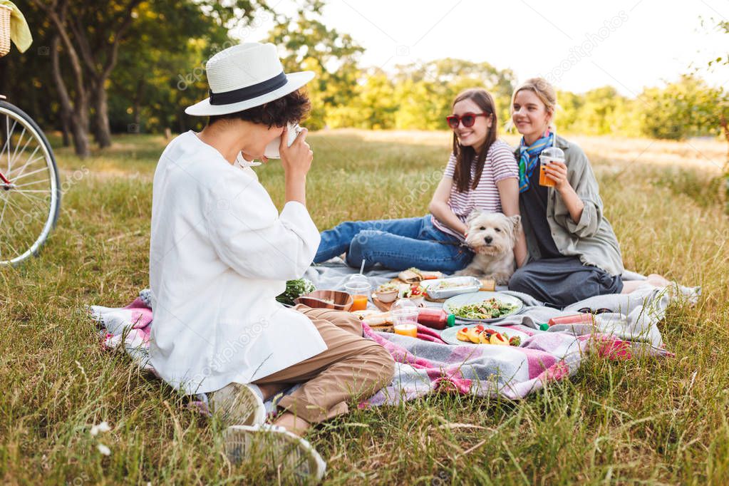 Two beautiful smiling girls sitting with little dog on picnic blanket and posing while girl in hat taking photos on camera on picnic in park