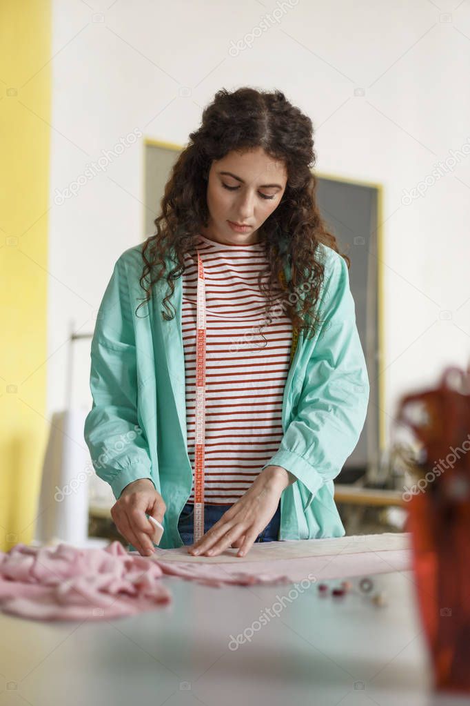 Beautiful designer of clothes in colorful shirt and striped T-shirt dreamily drawing with soap on pink textile in sewing workshop