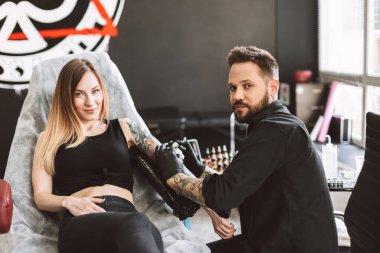 Tattooed girl and professional tattooer happily looking in camera while doing tattoo on hand by tattoo machine in studio clipart