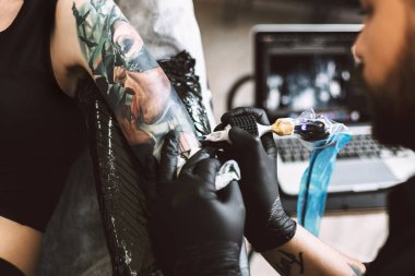 Close up professional tattoo master in black gloves doing tattoo on girl hand using tattoo machine in modern studio clipart