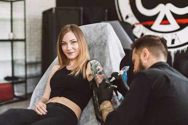 タトゥー スタジオ タトゥー マシンを使用しての手を行うプロの Tattooer ながらカメラで探しての刺青の女の子の笑顔 — ストック写真