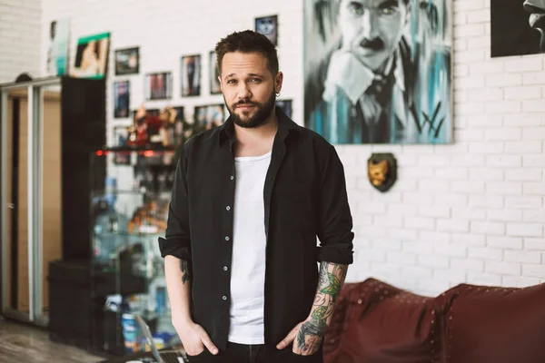 Professional tattooer standing in black shirt dreamily looking in camera spending time in modern tattoo studio