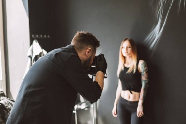 Dövmeli kız modern tattoo Studio siyah arka plan üzerinde ayakta, fotoğraf makinesinde fotoğrafların dövme ustası