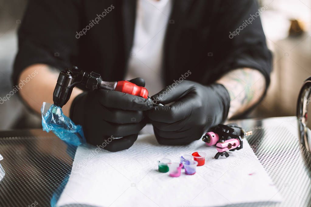 Close up tattooer in black gloves preparing tattoo machine for work with colorful inks in modern tattoo studio