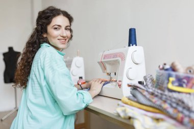 Renkli gömlek mutlu dikiş makinesi modern dikiş atölyesinde çalışan kamera seyir siyah kıvırcık saçlı gülümseyerek terzi