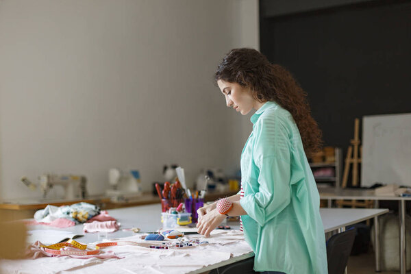 Молодая швея в рубашке вдумчиво выбирает красочную нить для работы с тканями в современной швейной мастерской
