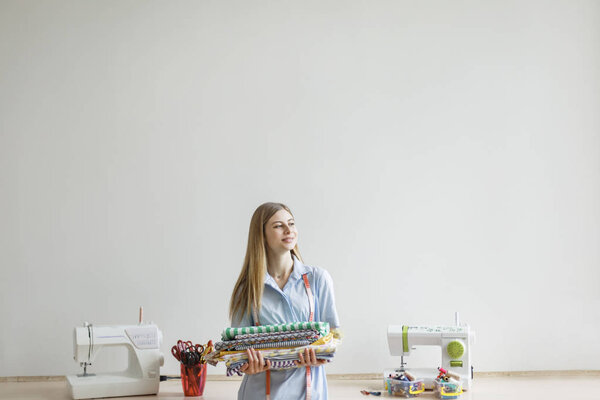 Young smiling seamstress in blue shirt joyfully looking aside holding patterns fabrics in hands in sewing workshop