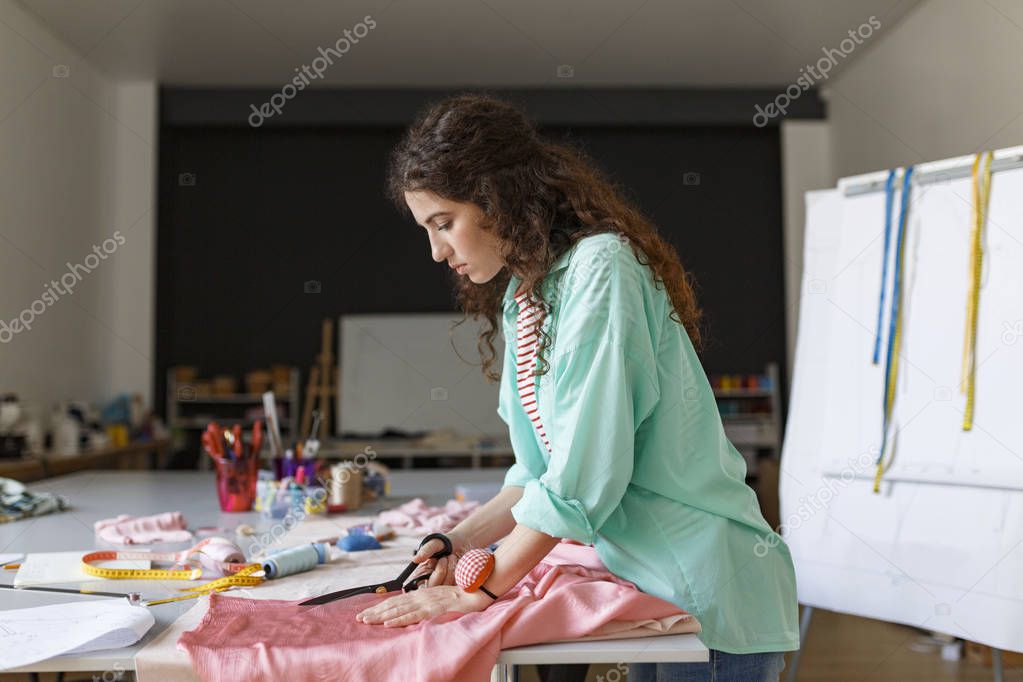 Young thoughtful girl in colorful shirt dreamily cutting fabric with scissors in modern sewing workshop