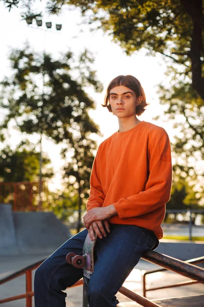 年轻的溜冰者在橙色套衫若有所思地看着在相机与滑板倚在栏杆上现代 Skatepark — 图库照片