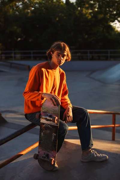 年轻的溜冰者在橙色套衫若有所思地看着在相机与滑板在 Skatepark — 图库照片
