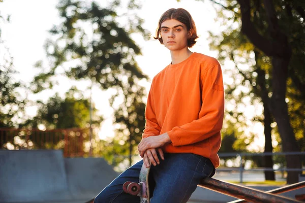 年轻严重的溜冰者在橙色套衫若有所思地看着在相机与滑板倚在栏杆上现代 Skatepark — 图库照片