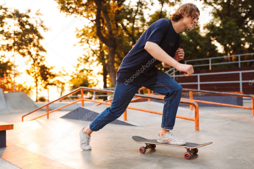 Young cool skater in black T-shirt and jeans riding on skateskateboard at skate park with sunrise on background