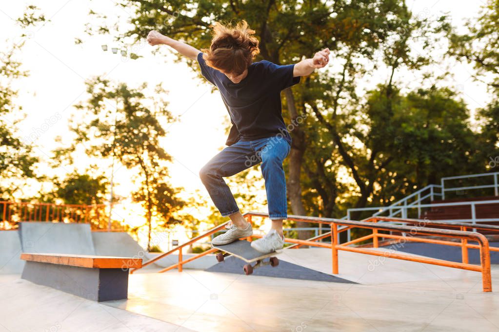 Young cool skater in black T-shirt and jeans practicing jumping tricks on skateboard at skate park