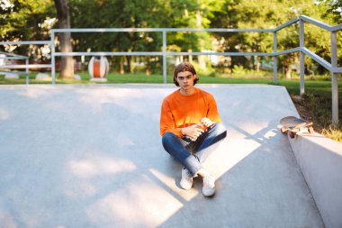 Genç adam turuncu kazak ve şişe suyu sersem kamera seyir skatepark elinde tutan kot
