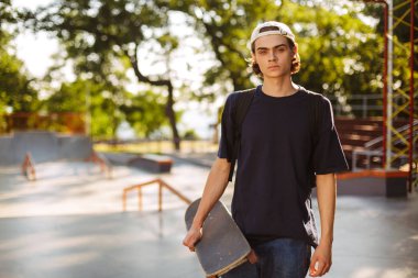 Genç ciddi adam siyah T-shirt ve beyaz kap düşünceli kamera kaykay el ile modern skatepark arka planda tutan arıyorsunuz