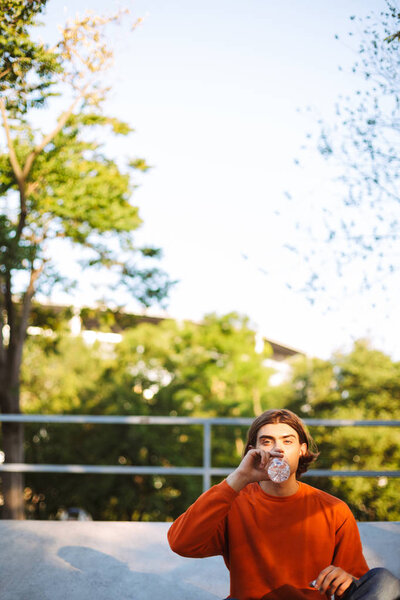 Young guy in orange pullover dreamily looking in camera drinking water at skatepark with beautiful view on background