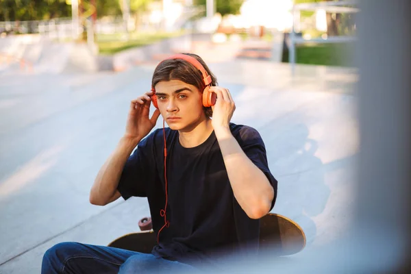 年轻的家伙在橙色耳机若有所思地看着相机听音乐与滑板在 Skatepark — 图库照片