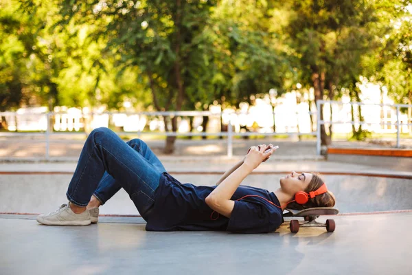 Νέοι Σκέιτερ Πορτοκαλί Ακουστικά Ξαπλωμένη Για Skateboard Σκεπτικά Χρησιμοποιώντας Κινητό — Φωτογραφία Αρχείου