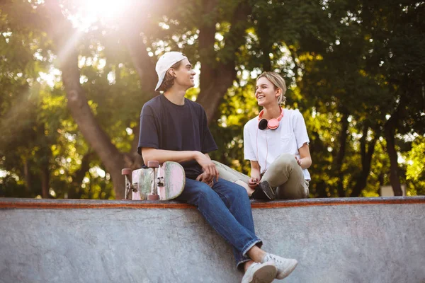 微笑的女孩与耳机和年轻的家伙与滑板快乐地花时间在一起在 Skatepark — 图库照片