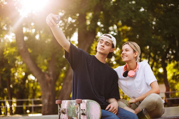 微笑的女孩与耳机和年轻人与滑板愉快地拍照在手机正面照相机在 Skatepark — 图库照片