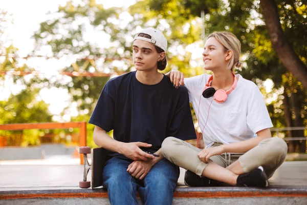 年轻的家伙与手机和漂亮的女孩与耳机高兴地看着一边花时间在 Skatepark — 图库照片