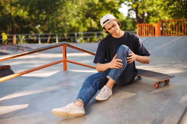 年轻的不安的溜冰者在盖帽举行他的痛苦的腿与滑板在 Skatepark — 图库照片