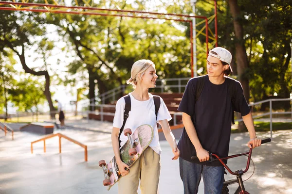 年轻的家伙与自行车和漂亮的微笑的女孩与滑板快乐地花时间在一起在现代 Skatepark — 图库照片