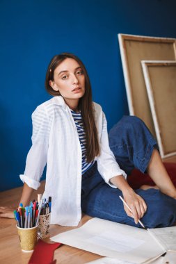 Beyaz gömlek ve çizgili tişört düşünceli kamera tuvaller ile evde arka plan üzerinde arıyorum elinde kalem tutan katta oturan dalgın ressam