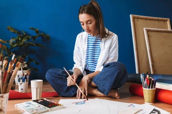 Pensive girl in white shirt and striped T-shirt sitting on floor dreamily drawing by watercolor spending time at home