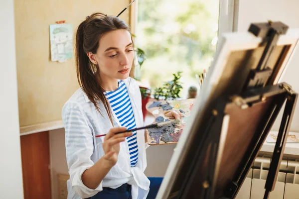 Thoughtful creative girl in white shirt and striped T-shirt holding paint brush in hand dreamily drawing on easel at home