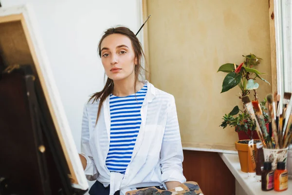 stock image Beautiful girl in white shirt and striped T-shirt dreamily looking in camera drawing on easel with paint tools near on window sill at home