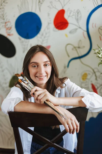 Cheerful girl in white shirt on chair joyfully looking in camera holding paint brushes in hand with big patterns painting on background at home