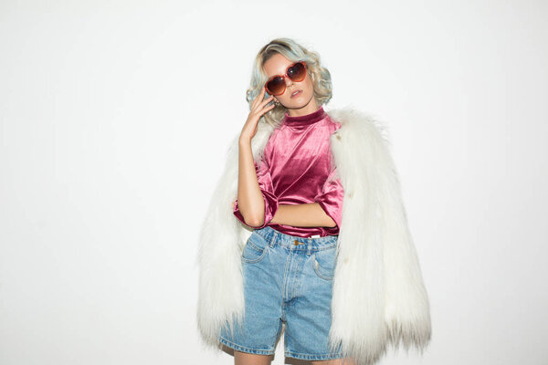 Beautiful fashion girl in white fur jacket and sunglasses dreamily looking in camera over white background 