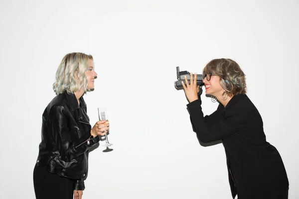 Beautiful smiling girl in black jacket happily taking photos on polaroid camera of pretty girl with glass of champagne in hand over white background