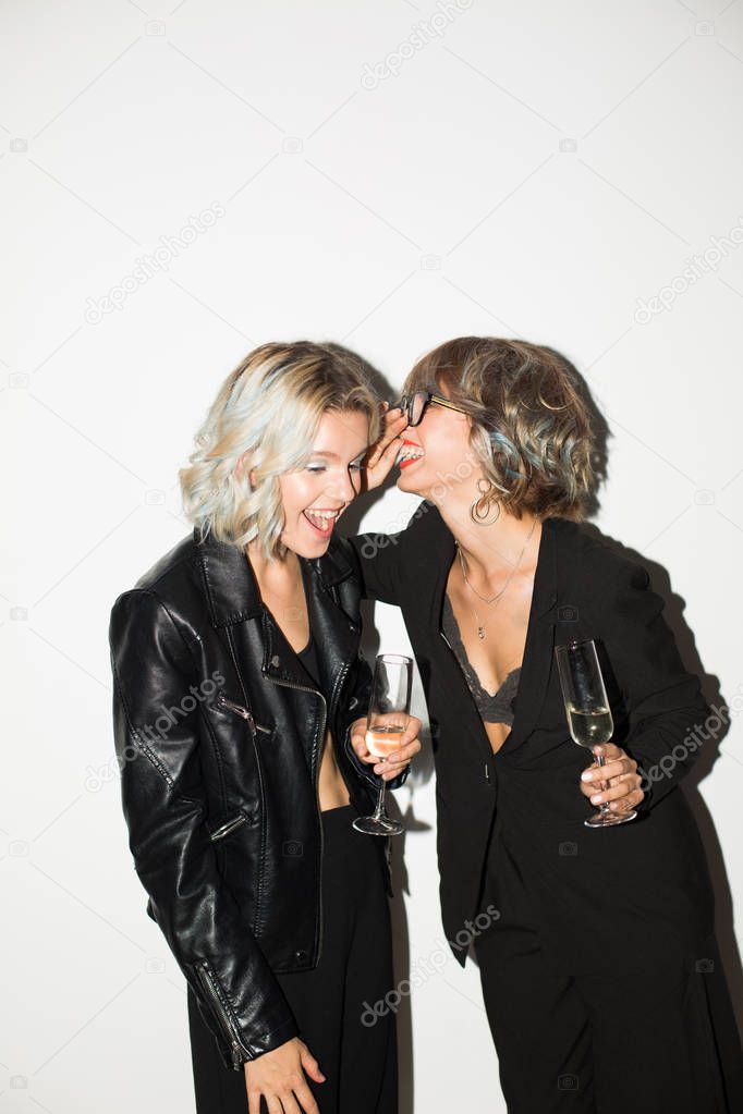 Two beautiful smiling girls in black jackets with glasses of champagne in hands happily gossiping over white background