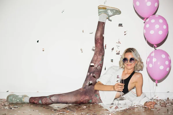 Pretty smiling girl in sunglasses holding glass of champagne and pink balloons in hands happily looking in camera lifting leg up over white background