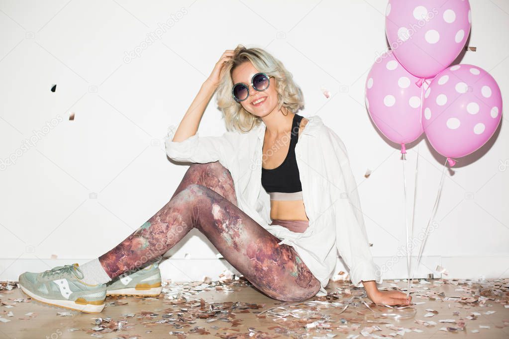 Pretty smiling girl in sunglasses with pink balloons happily looking in camera with confetti on floor over white background