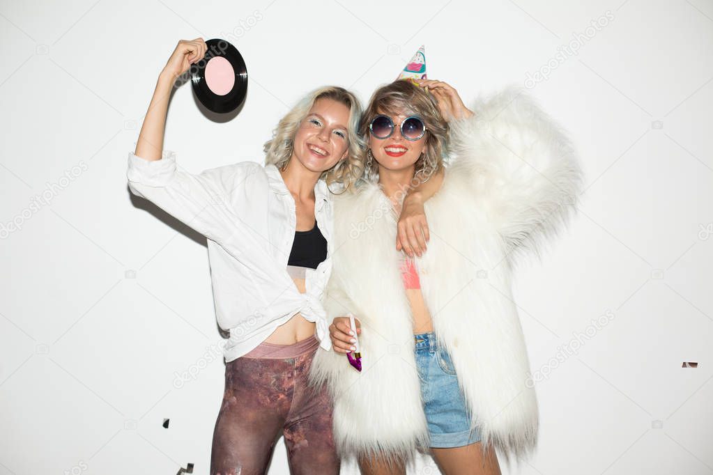 Beautiful girl in birthday cap and white fur jacket and pretty girl in shirt near holding retro music plate and happily looking in camera together over white background