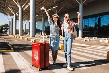 Two cheerful female friends in sunglasses happily looking in camera raising hands up with red suitcase and backpack on shoulder outdoor near airport clipart