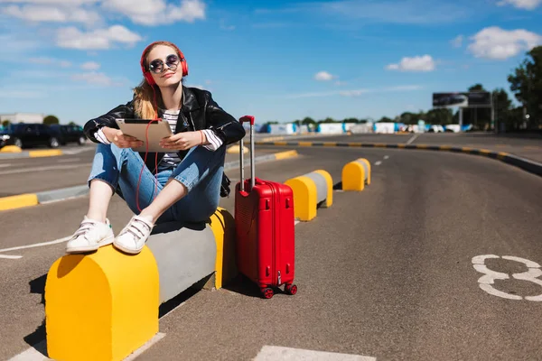 Güzel kız güneş gözlüğü ve tablet yolda Havaalanı kırmızı çanta ile müzik dinlerken kulaklık