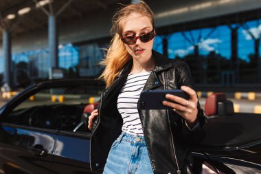 Güneş gözlüğü ve deri ceket cep telefonu arka plan siyah cabriolet araba ile fotoğraf çekmek güzel kız