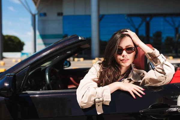 Beautiful girl in sunglasses and trench coat leaning on cabriolet car door dreamily looking in camera with airport on background