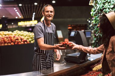 Önlük mutlu müşteri modern süpermarkette kamera vererek çilek isteyen tezgahın arkasındaki genç gülümseyen satıcısı