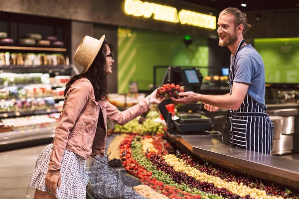 年轻微笑卖家在围裙后面柜台愉快地给顾客在现代超级市场的草莓 — 图库照片