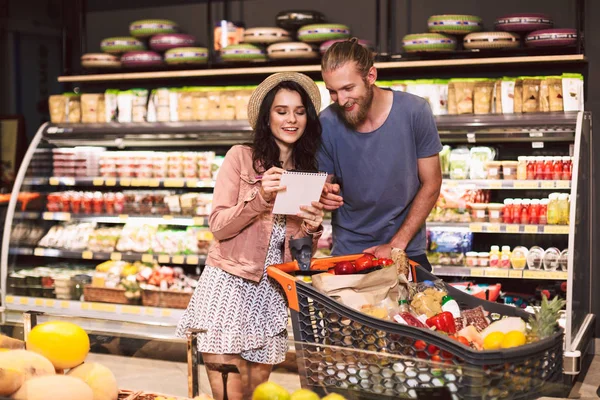 年轻夫妇与手推车充满产品阅读购物单花时间一起在现代超市 — 图库照片