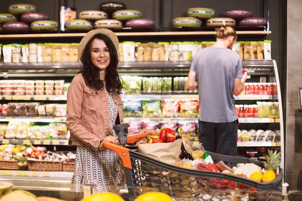Beautiful smiling girl in hat happily looking in camera with trolley full of products while guy on background choosing products in modern supermarket