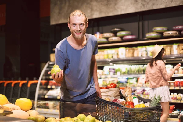 Young smiling man joyfully looking in camera holding pear in hand with trolley full of products near while pretty girl on background choosing products in modern supermarket